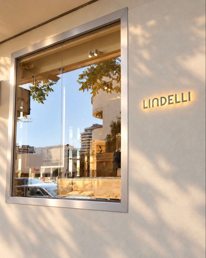 A jewellery storefront exterior with a large rectangular window, a white wall, and the brand name LINDELLI in illuminated gold lettering on the wall. A smaller LINDELLI sign is visible on the left, and reflections of trees and buildings appear in the glass.